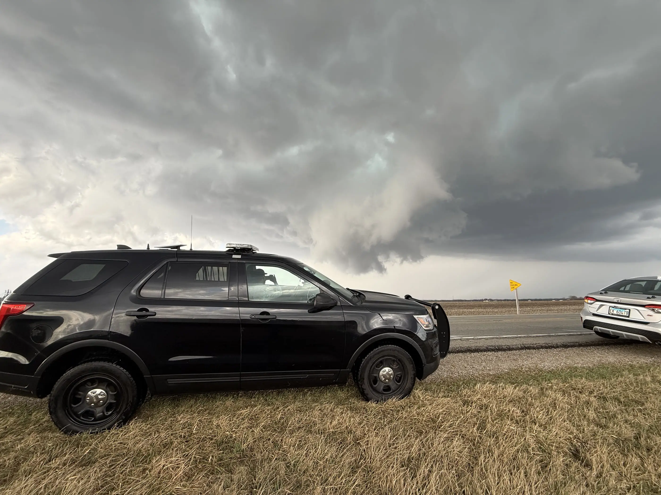The chase vehicle parked on a grass shoulder, a lowering storm base and rain shaft behind it.