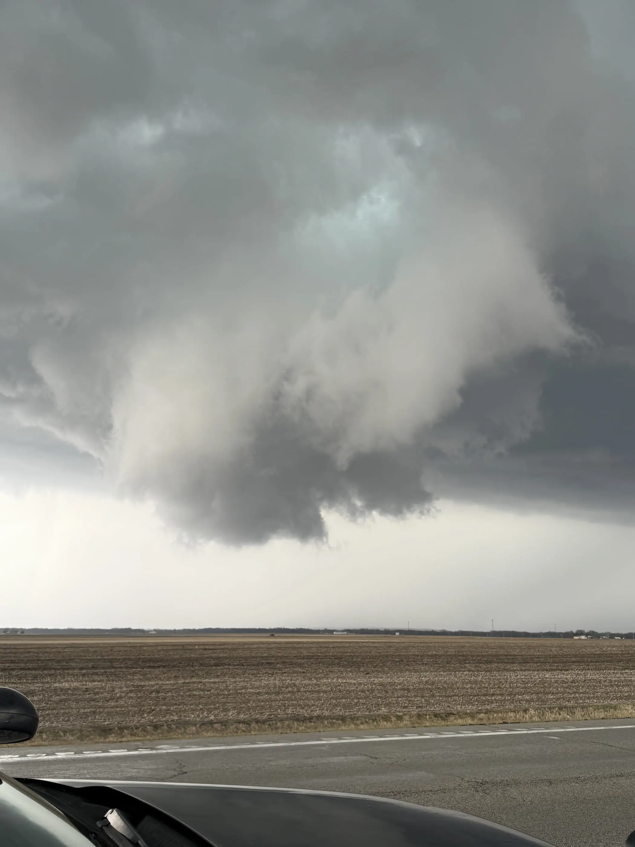 A wall cloud and rain-free base above open farmland, shot over the hood of the chase vehicle.
